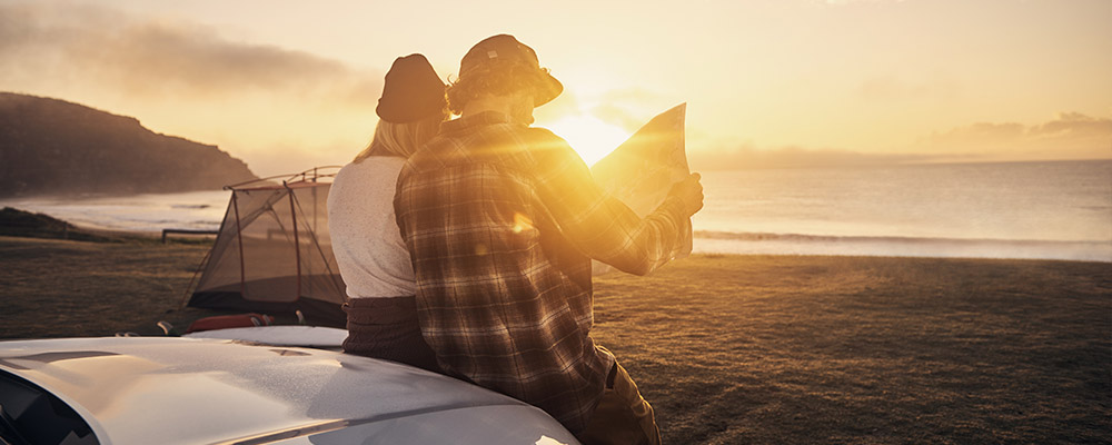 Couple looking at a map, sitting on the bonnet of a car in front of the ocean